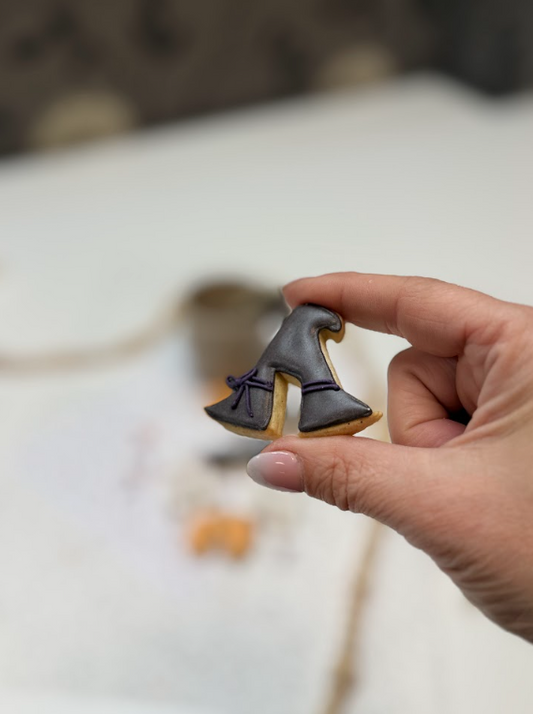 Hand houdt een heksenhoedkoekje vast, gedecoreerd met donkerpaarse en zwarte royal icing.