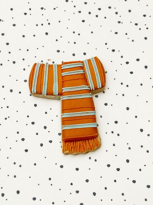 Koekje in de vorm van een oranje sjaal met witte en lichtblauwe strepen, op een gestippelde ondergrond.