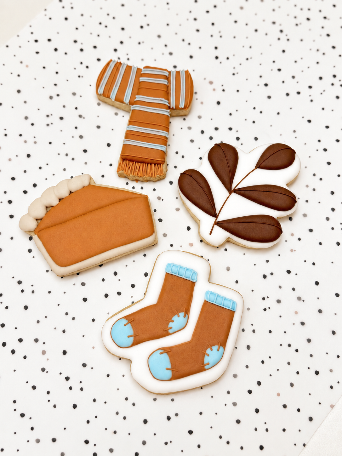 Vier herfstkoekjes op een gestippelde ondergrond: een sjaal, een taartpunt, een blad en een paar sokken, versierd met royal icing.