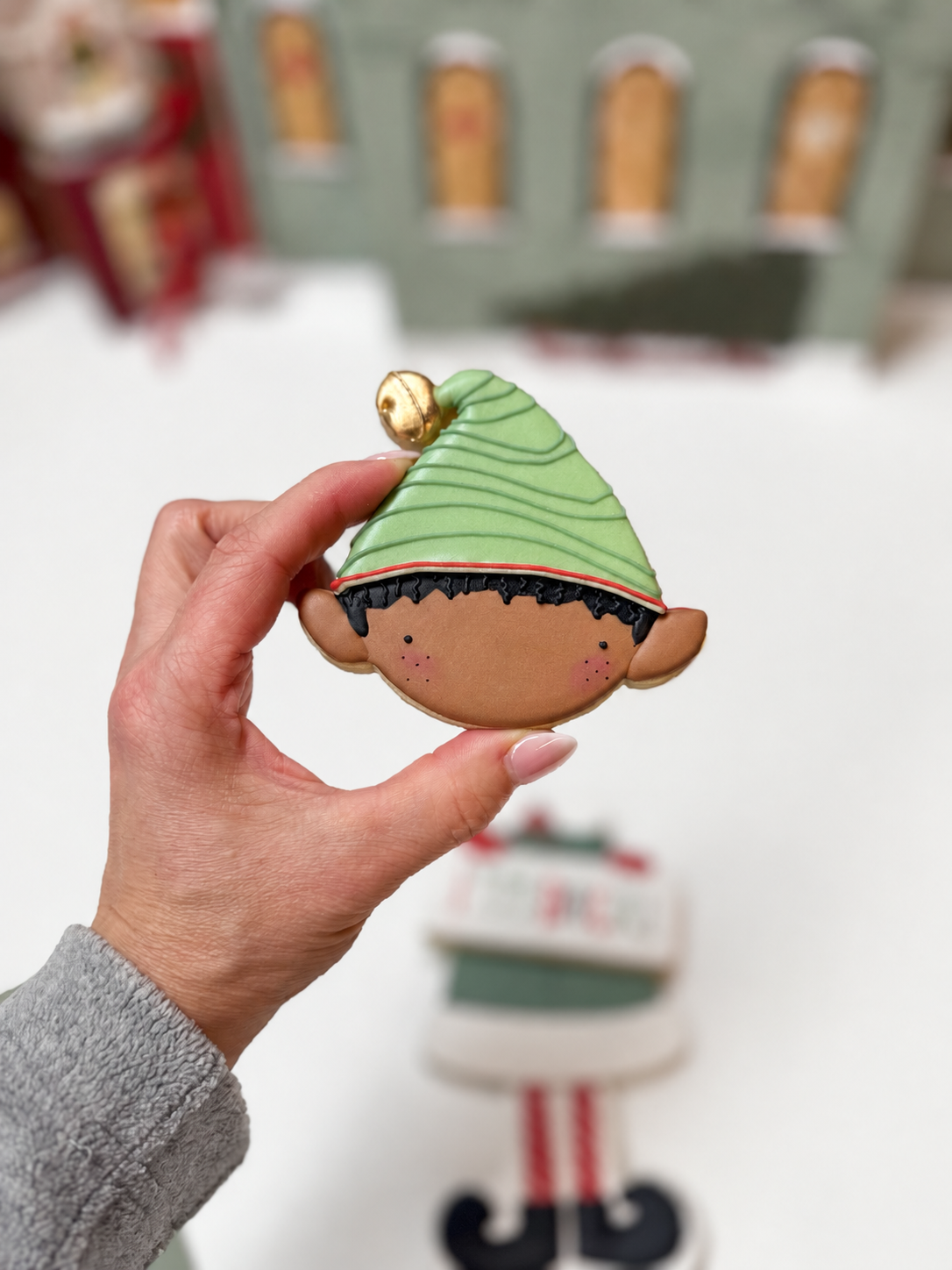 Hand houdt een kerstelfkoekje met donker haar, een groen gestreepte muts en een gouden belletje vast. De overige elfkoekjes liggen op de achtergrond.