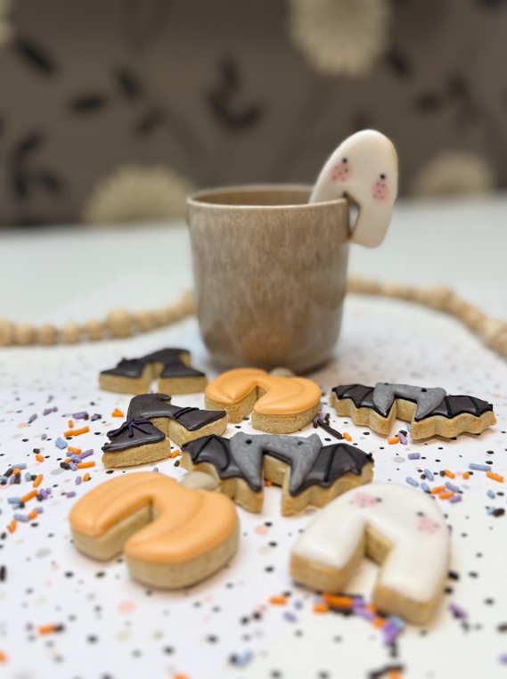 Halloween koekjes in de vorm van vleermuizen, pompoenen en spookjes, naast een mok met een spookkoekje aan de rand.