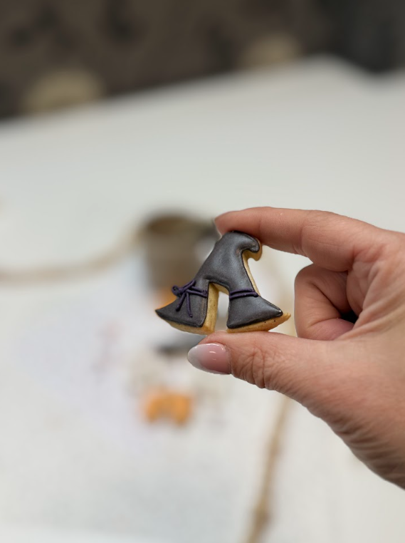 Hand houdt een heksenhoedkoekje vast, gedecoreerd met donkerpaarse en zwarte royal icing.