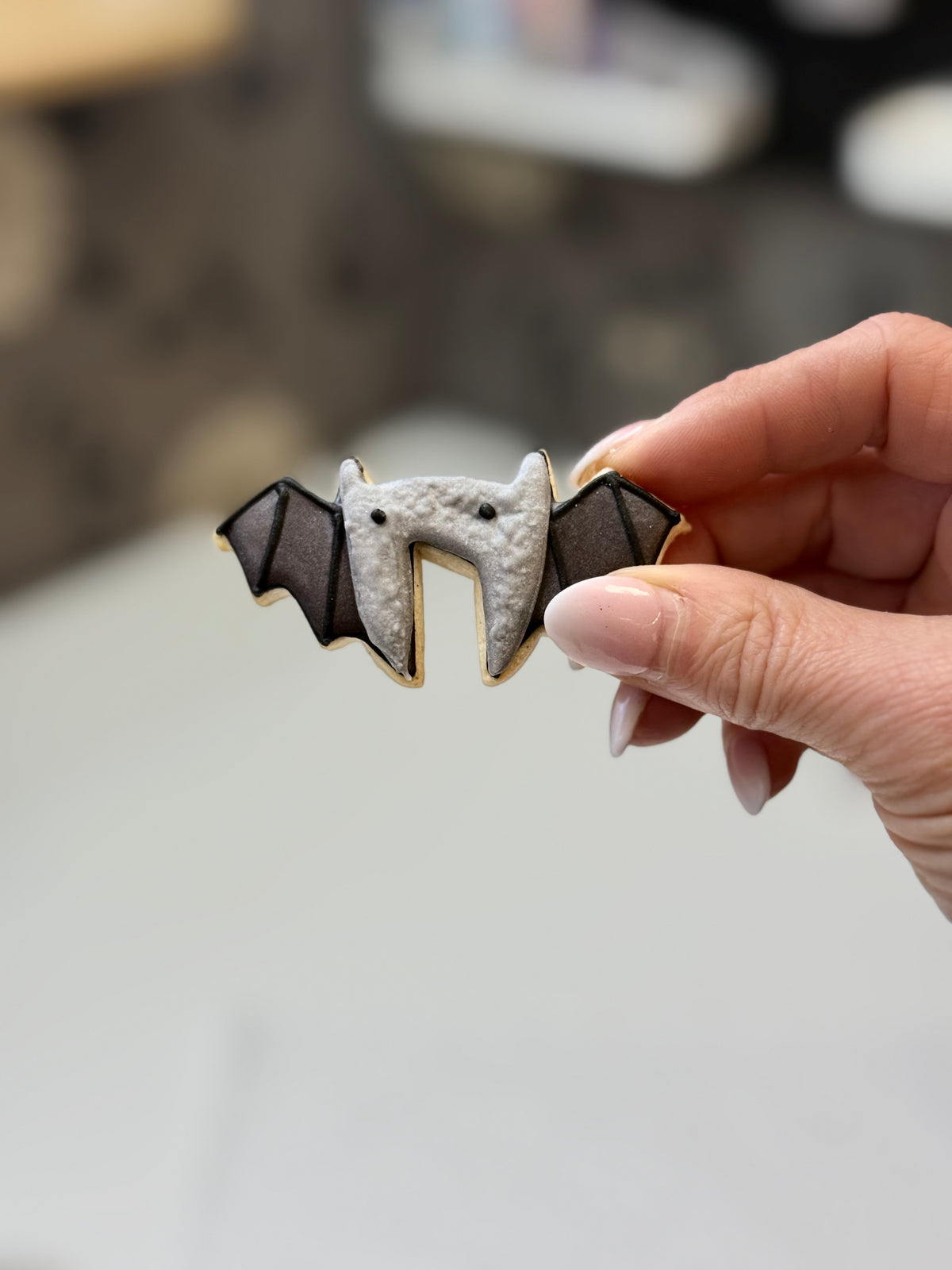Hand houdt een vleermuiskoekje vast met een grijs lijfje, zwarte oogjes en donkere vleugels.