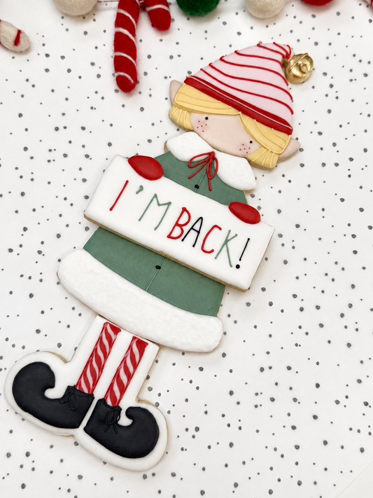 Kerstelf gemaakt van vier gedecoreerde koekjes, met blond haar, een rood-wit gestreepte muts, groene jas en een bordje met ‘I’m Back!’.