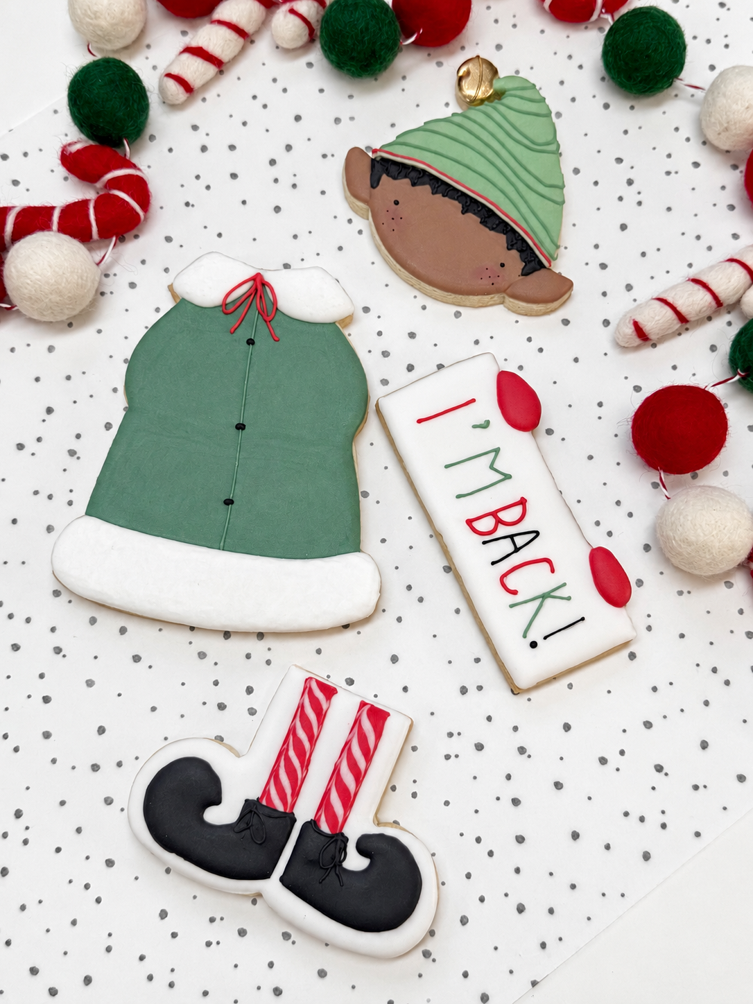 Vier losse kerstelfkoekjes: een hoofd met donker haar en groene muts, een groene jas, een bordje ‘I’m Back!’ en rood-wit gestreepte benen met zwarte schoenen, op een gestippelde ondergrond.