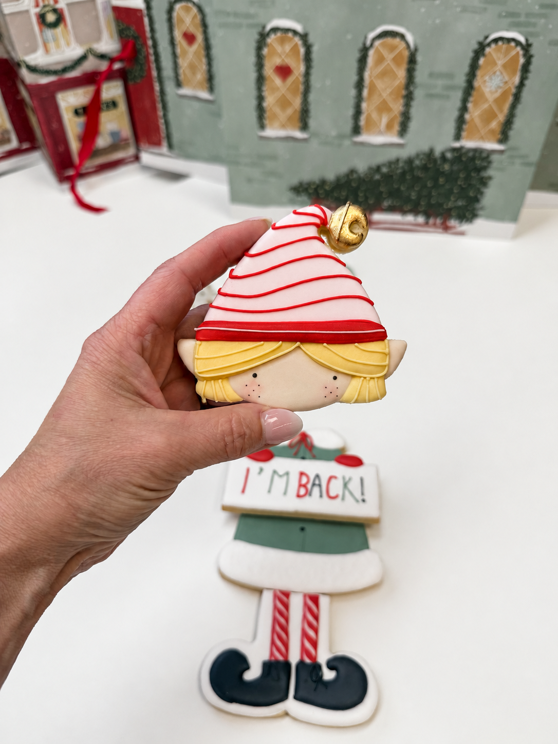 Hand houdt een kerstelfkoekje met blond haar, een rood-wit gestreepte muts en een gouden belletje vast. De overige elfkoekjes liggen op de achtergrond.