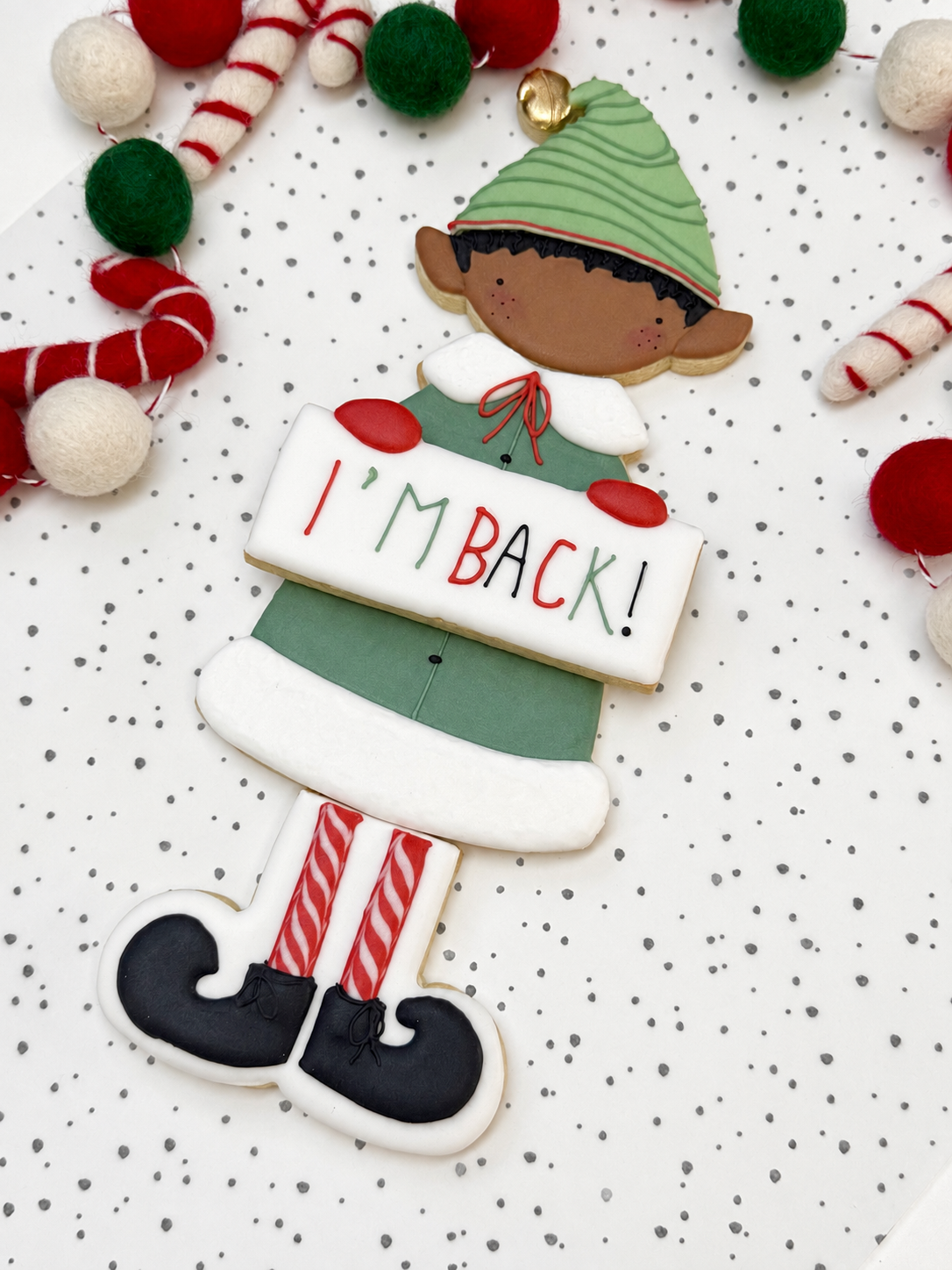 Kerstelf van vier gedecoreerde koekjes met donker haar, een groen gestreepte muts, groene jas, bordje ‘I’m Back!’ en rood-wit gestreepte benen met zwarte schoenen.